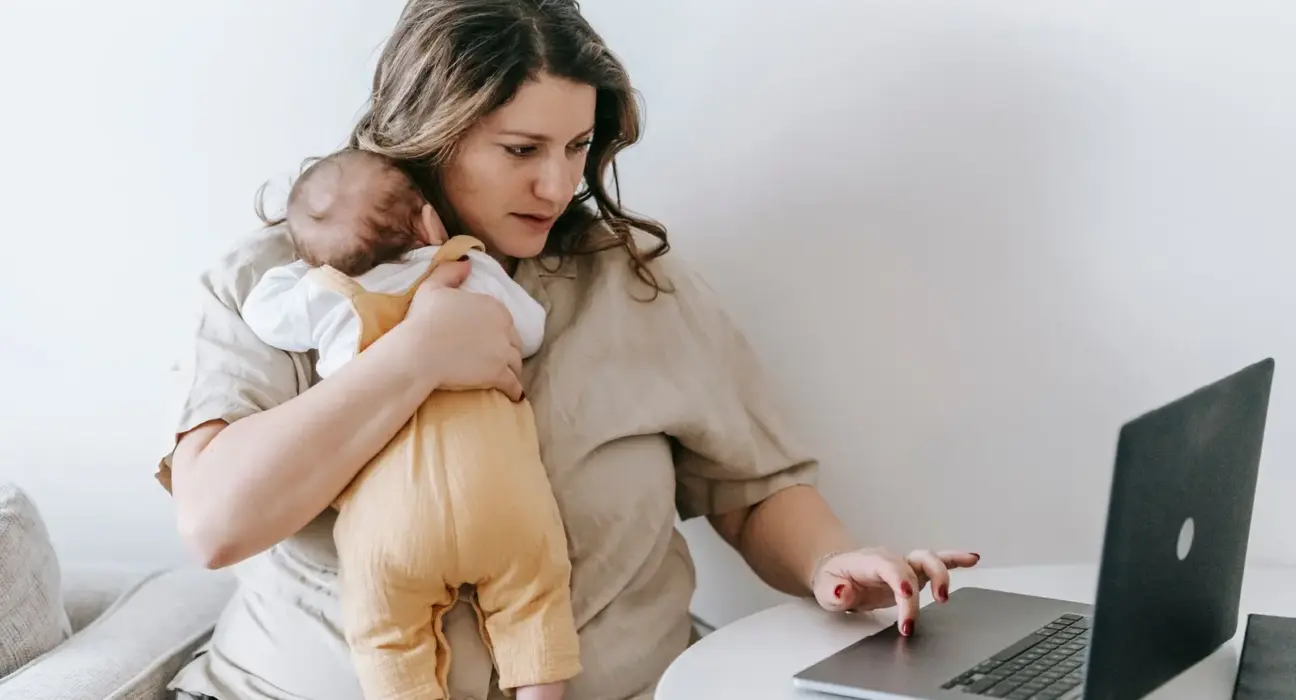 Selbstständige Mutter arbeitet am Laptop und hält ihr Baby im Arm