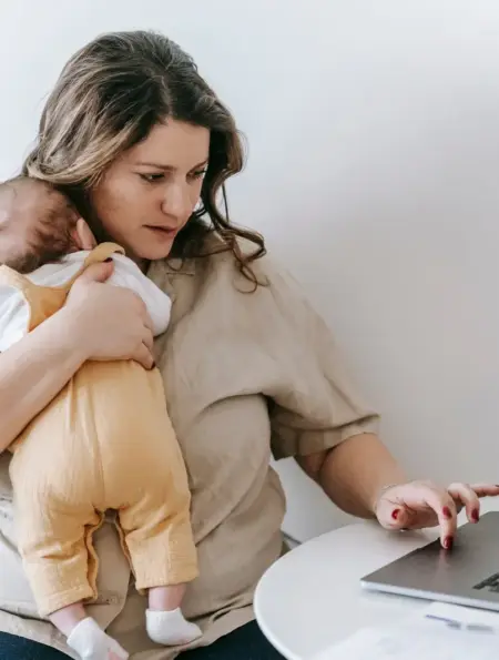 Selbstständige Mutter arbeitet am Laptop und hält ihr Baby im Arm