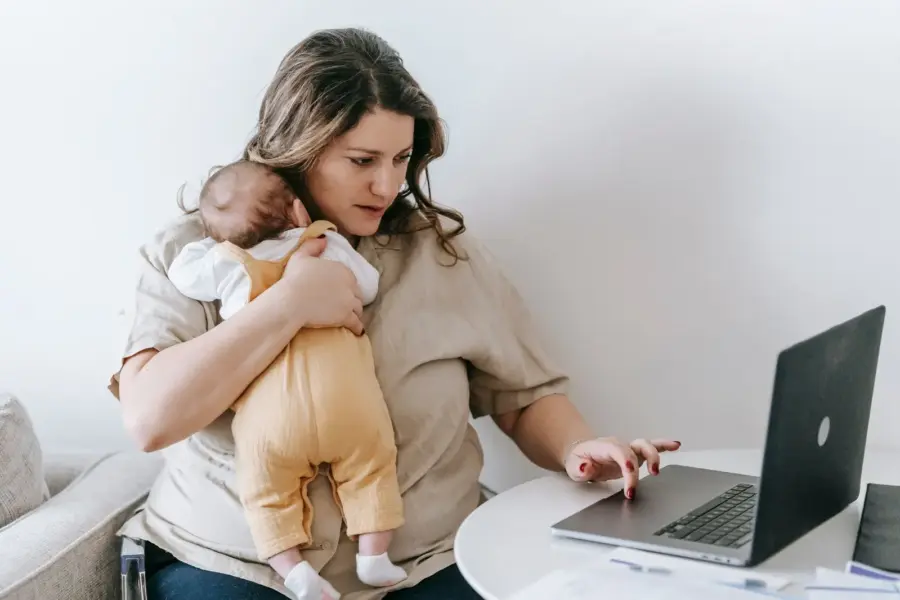 Selbstständige Mutter arbeitet am Laptop und hält ihr Baby im Arm