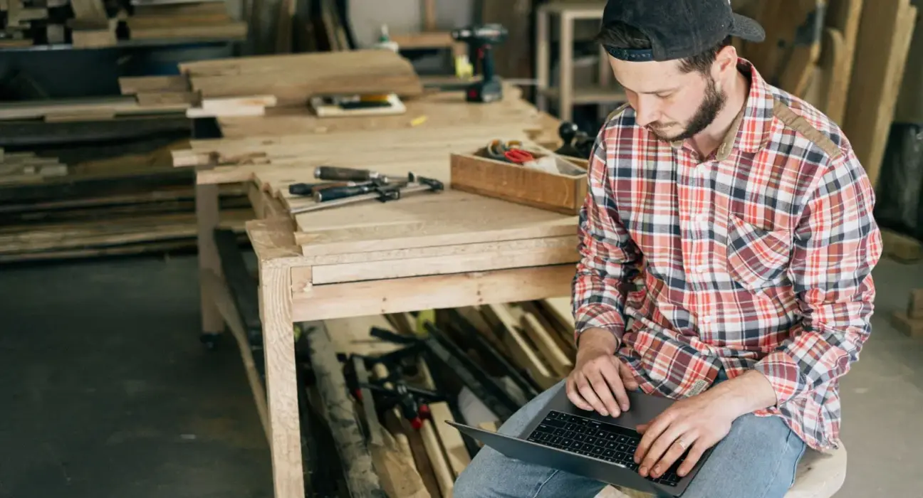 Handwerker plant am Laptop zwischen Werkzeugen und Werkbänken in seiner Werkstatt