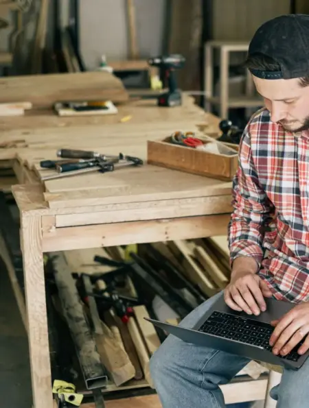 Handwerker plant am Laptop zwischen Werkzeugen und Werkbänken in seiner Werkstatt