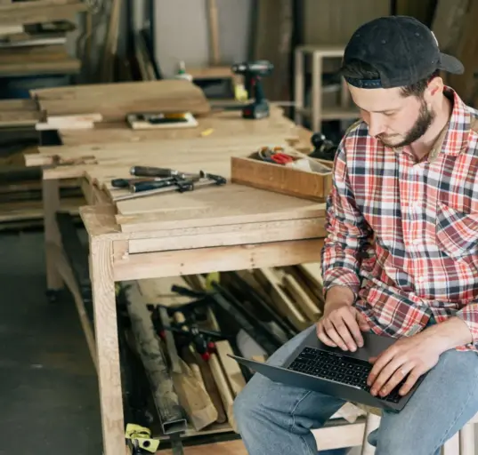 Handwerker plant am Laptop zwischen Werkzeugen und Werkbänken in seiner Werkstatt