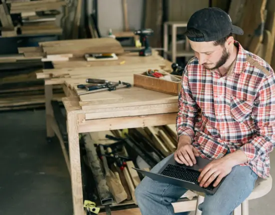 Handwerker plant am Laptop zwischen Werkzeugen und Werkbänken in seiner Werkstatt