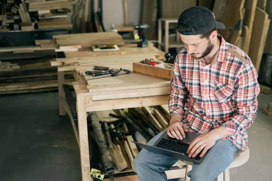 Handwerker plant am Laptop zwischen Werkzeugen und Werkbänken in seiner Werkstatt